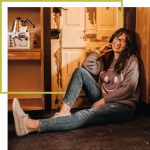 integrative business coach kristen king sitting on the floor against an old wooden door in jeans and a sweater with a smiley face on it